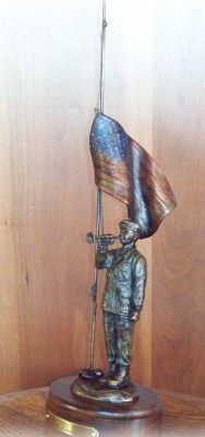 Bronze sculpture of soldier playing taps with half-mast American flag.