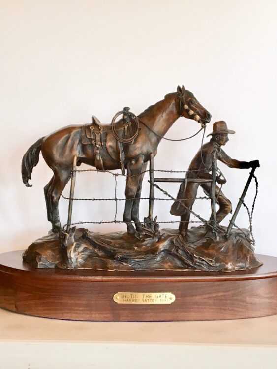 A cowboy with his saddle horse is shutting a barbed-wire gate.