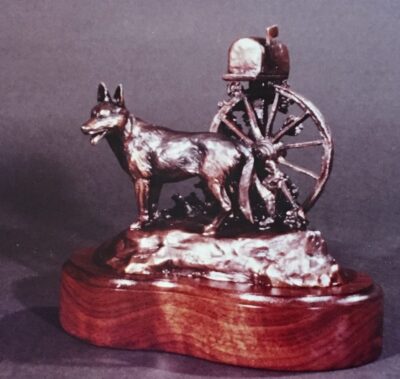 Bronze sculpture of German Shepard dog waiting by the mailbox that is mounted on a wagon wheel.