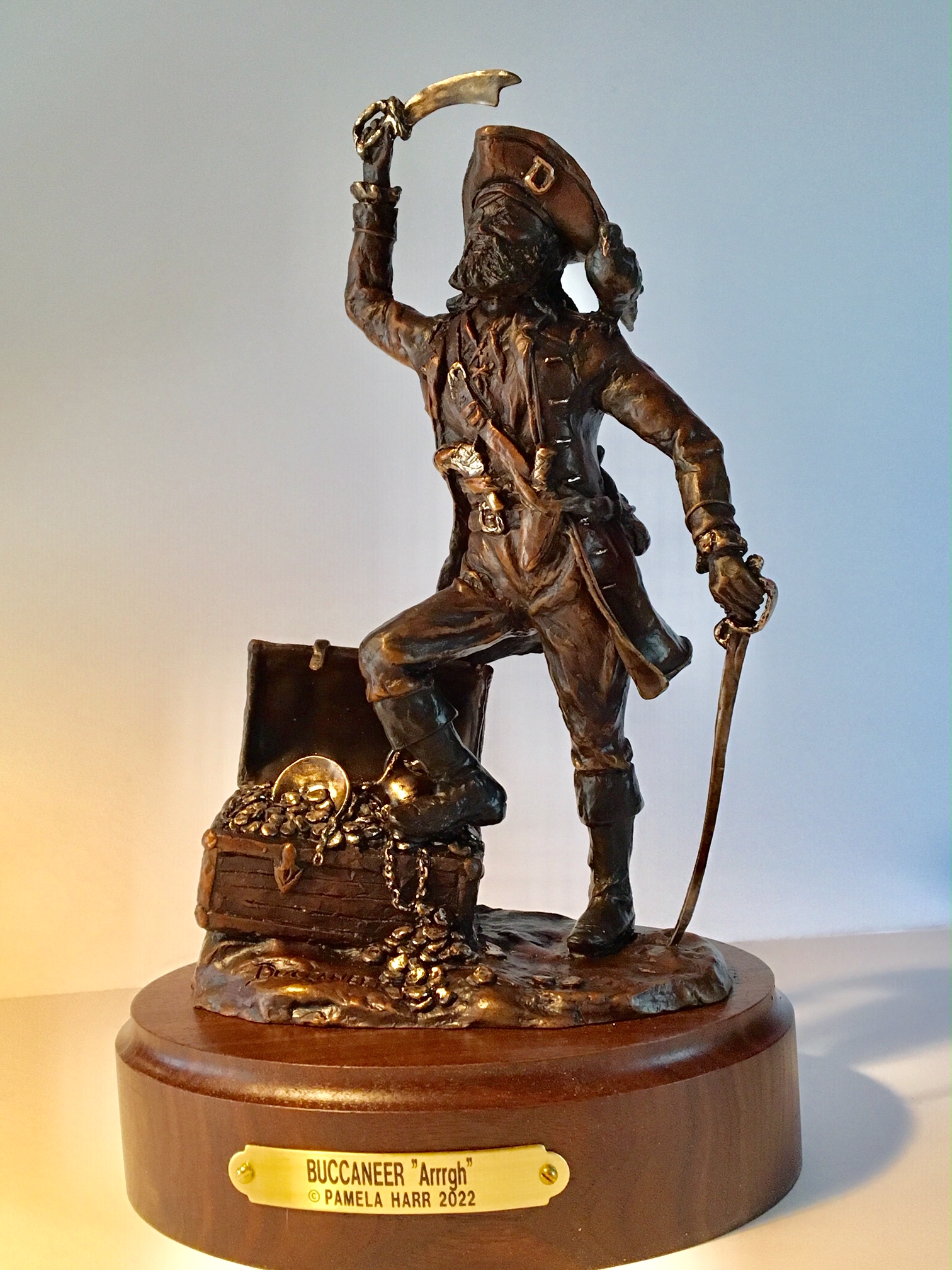 Bronze sculpture of a buccaneer with his foot on a treasure chest and a sword in his raised right hand.