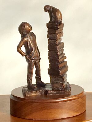 Bronze sculpture of a stack of books with a cat sitting on top of the books. Young child is looking up at the cat.