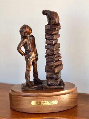 Charming bronze sculpture of a young girl looking at a cat who is sitting on top of a large stack of library books.