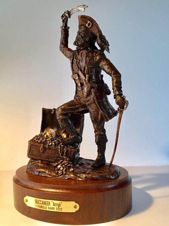 Bronze sculpture of a buccaneer with one foot propped on an overflowing treasure chest and a sword in each hand.