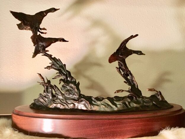 Bronze sculpture of three wild ducks preparing to land on choppy water near the cattails.
